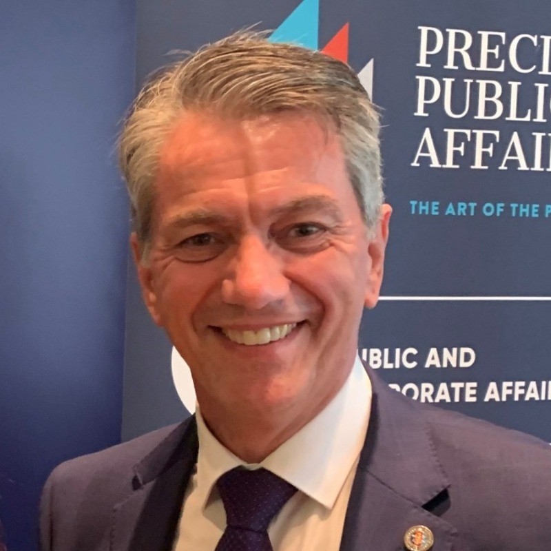 A smiling man in a blue suit and tie stands in front of a sign that partially reads "Precinct Public Affairs." The background features blue and white colors with some red accents.