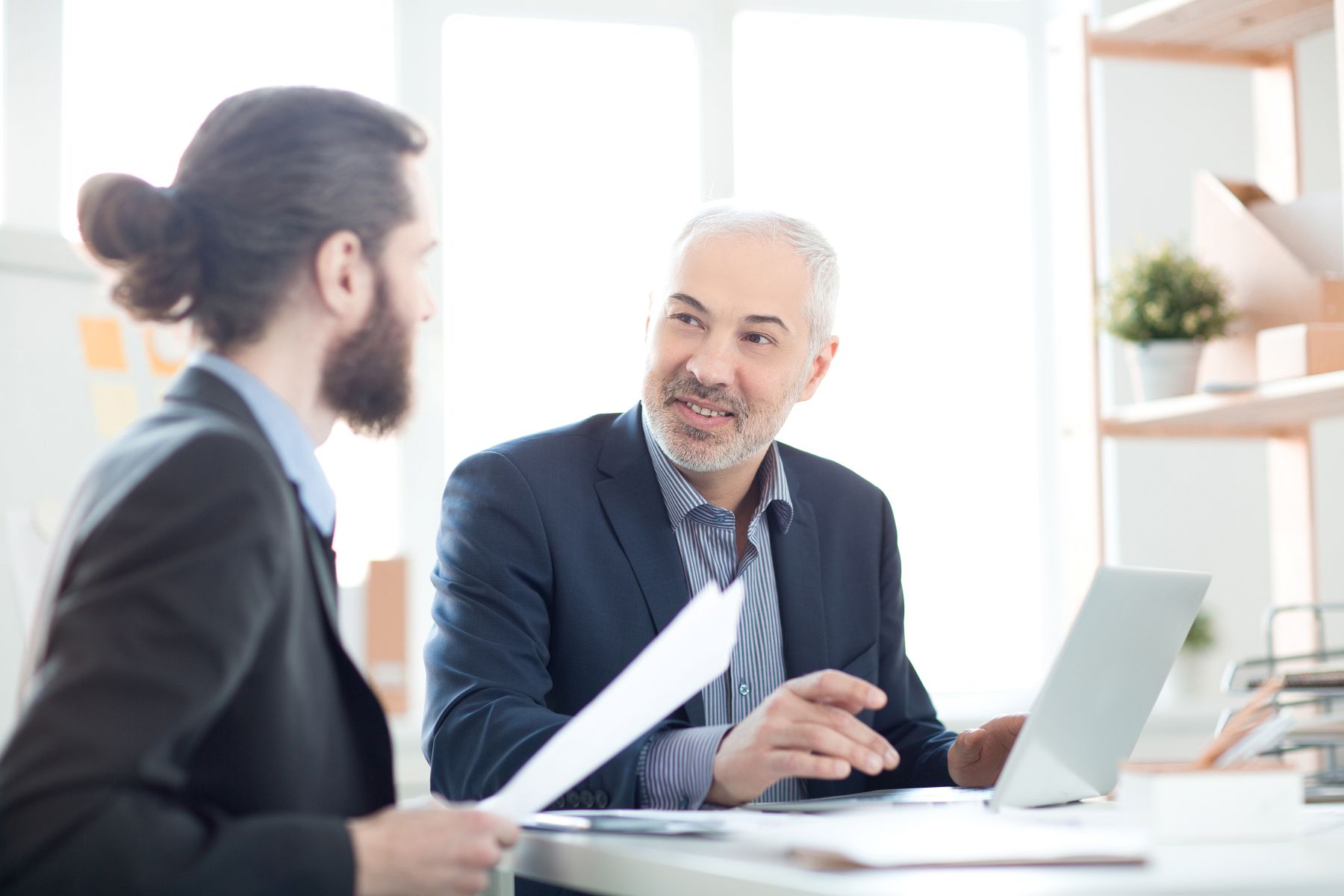 Two businessmen in suits are having a conversation in a bright office. One is holding papers, the other is using a laptop and smiling. Shelves and a plant are visible in the background.