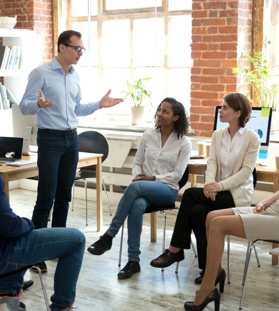 A man stands and speaks to three seated women in an office with brick walls and large windows. The group appears engaged in discussion, with laptops and plants in the background.