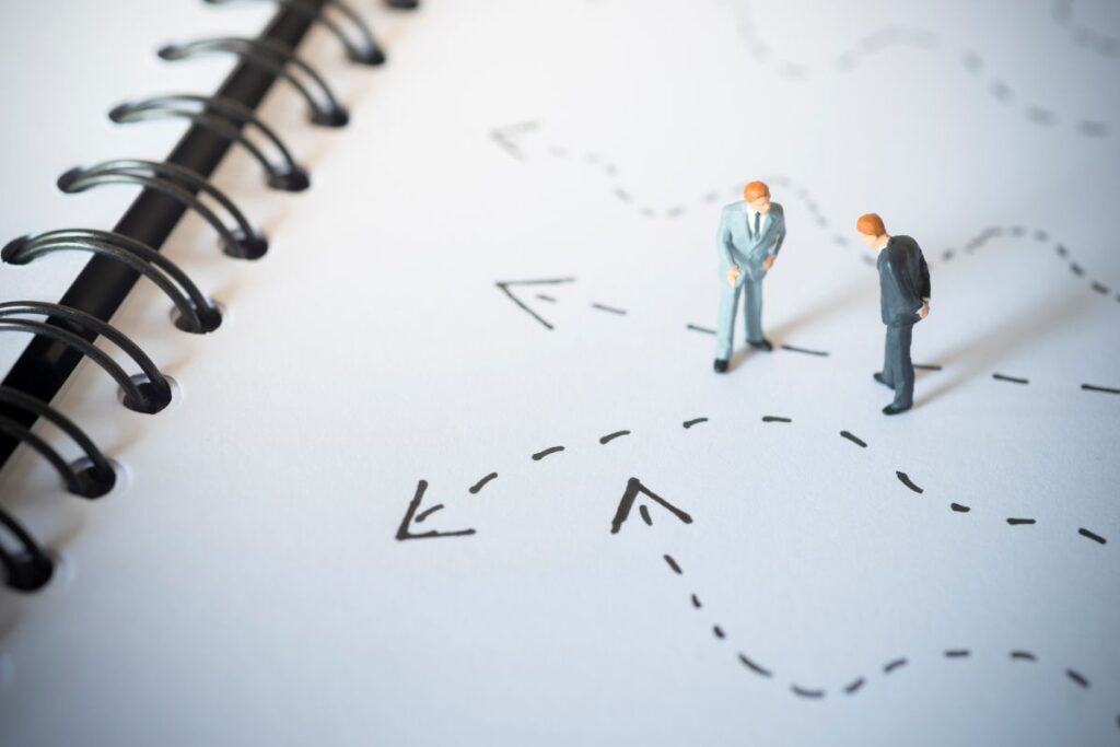 Two miniature business figures stand on a notebook page with dotted lines and arrows pointing in different directions, symbolizing decision-making and various paths.