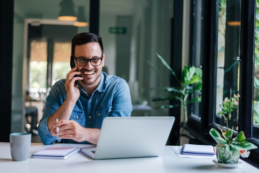A smiling man wearing glasses and a denim shirt sits at a desk with a laptop, notebook, and coffee mug, talking on the phone in a bright, modern office with large windows and green plants.