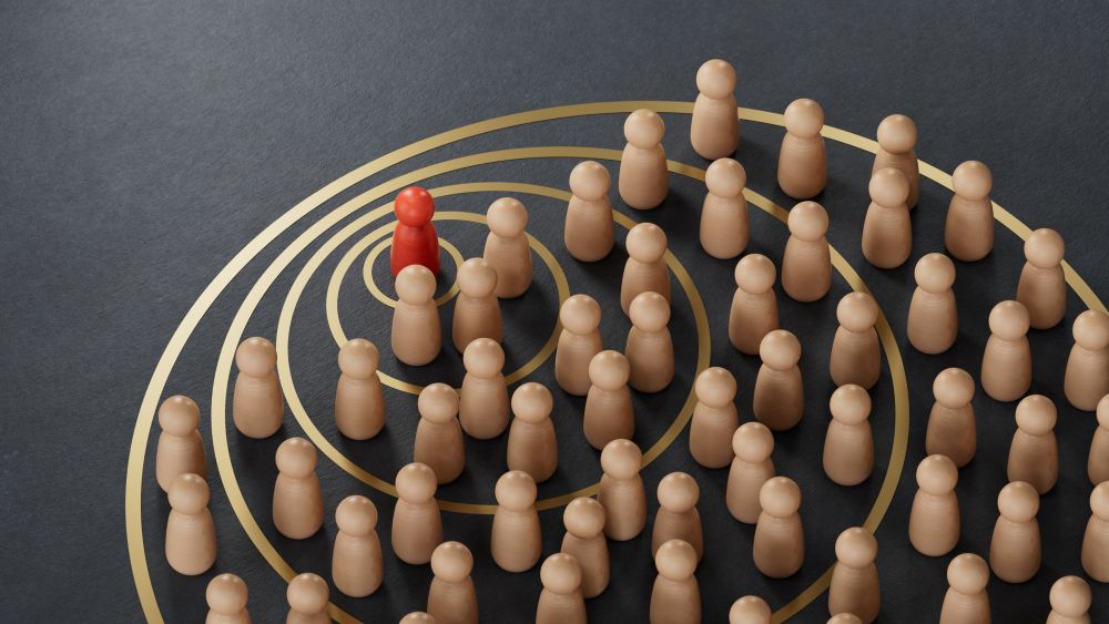 Several beige wooden figurines stand on black surface with gold concentric circles, surrounding a single red figurine positioned near the center, symbolizing individuality or leadership among a crowd.