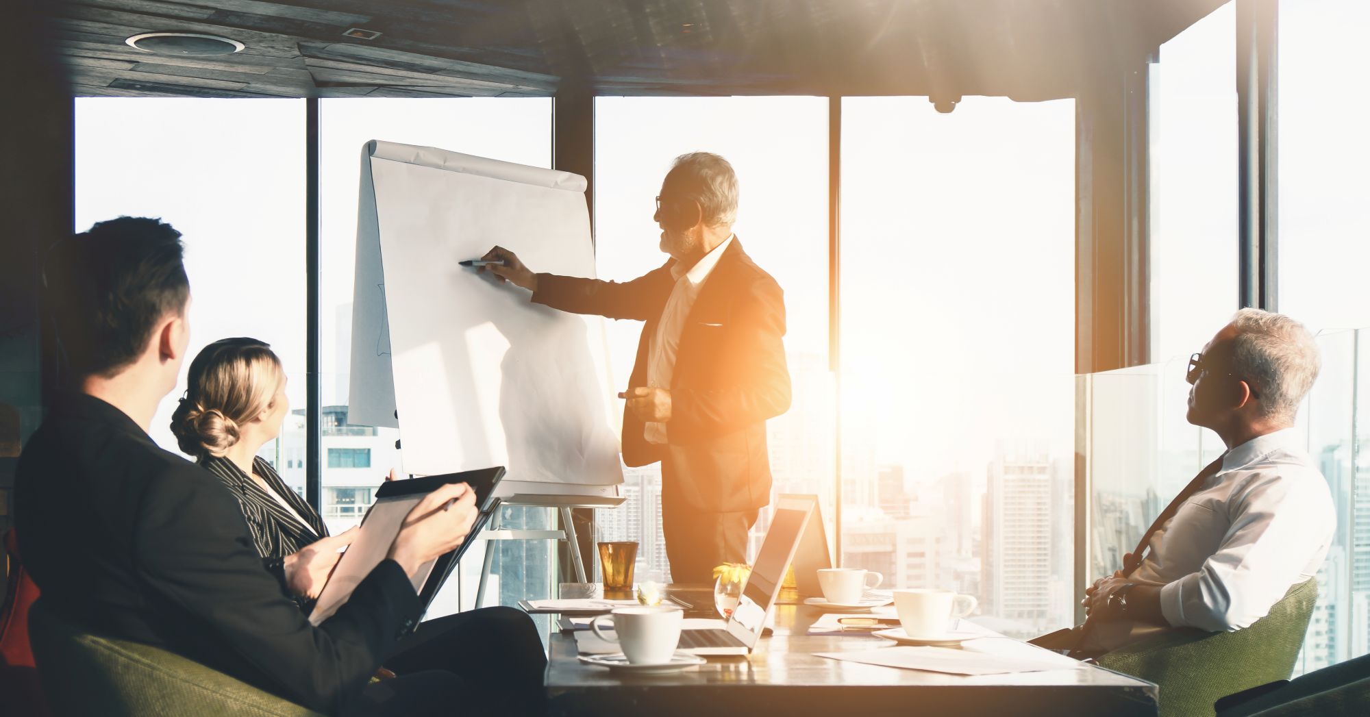 A business professional stands at a flip chart giving a presentation to three colleagues seated around a conference table in a modern office with large windows and a cityscape view.