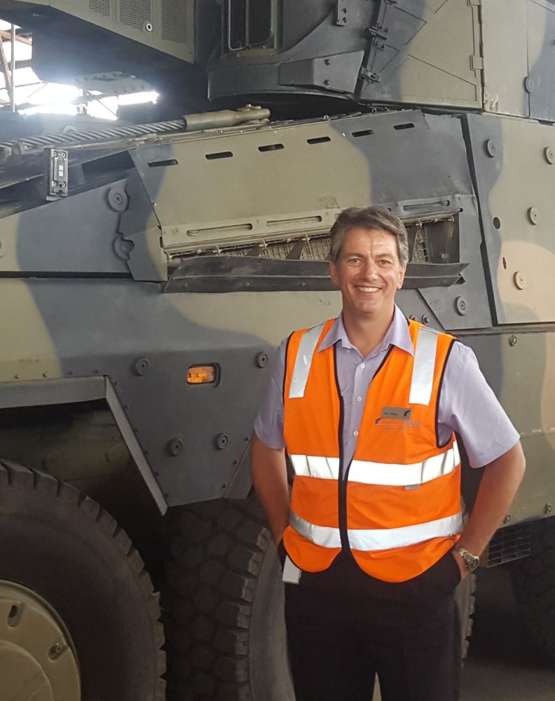 A man wearing a bright orange safety vest stands and smiles in front of a large military vehicle with a camouflage pattern inside an industrial setting.