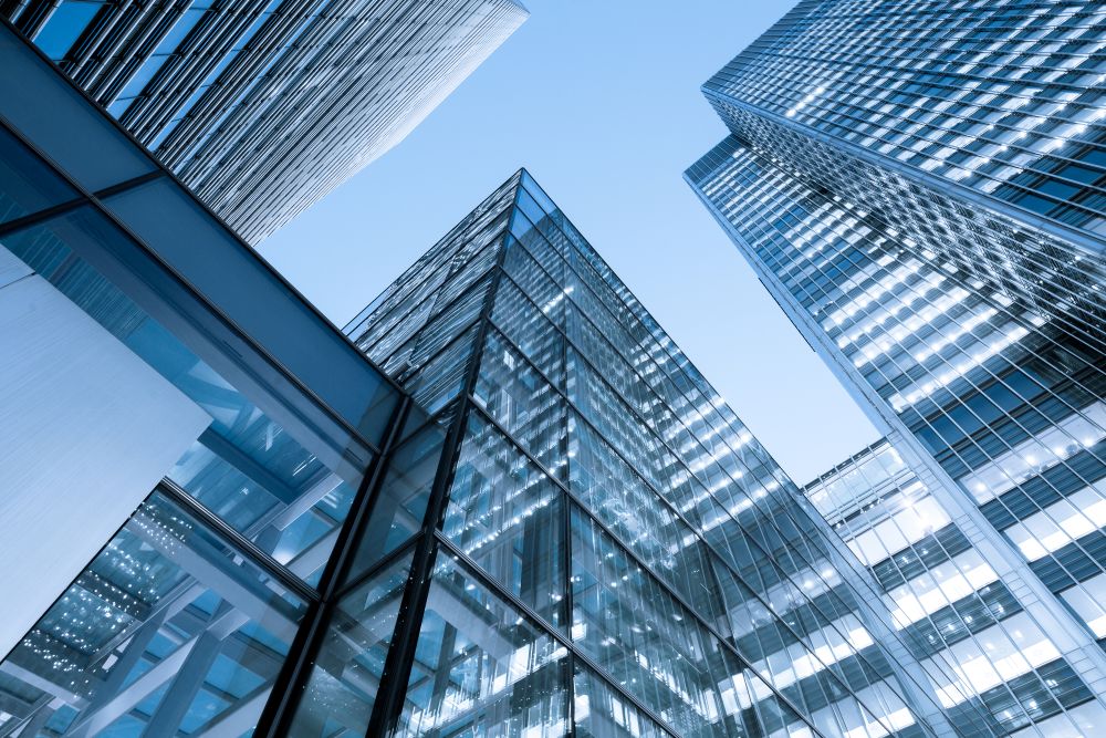 Tall, modern glass skyscrapers viewed from below against a clear blue sky, with reflections on the windows and geometric patterns created by the building facades.