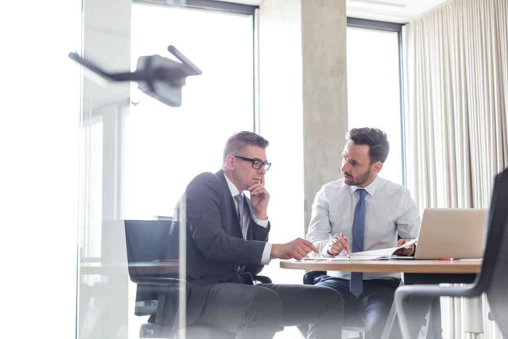Two businessmen in formal attire sit at a desk in a modern office, engaged in a serious discussion, with documents and a laptop open in front of them and large windows in the background.