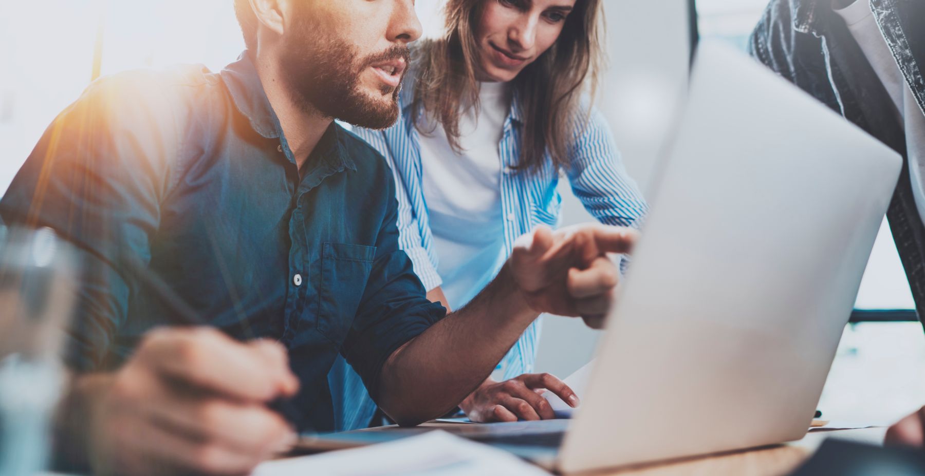 Two people are looking at a laptop screen together. One is pointing at the screen while the other listens and holds a pencil. Papers are on the table, suggesting they are collaborating or working on a project.