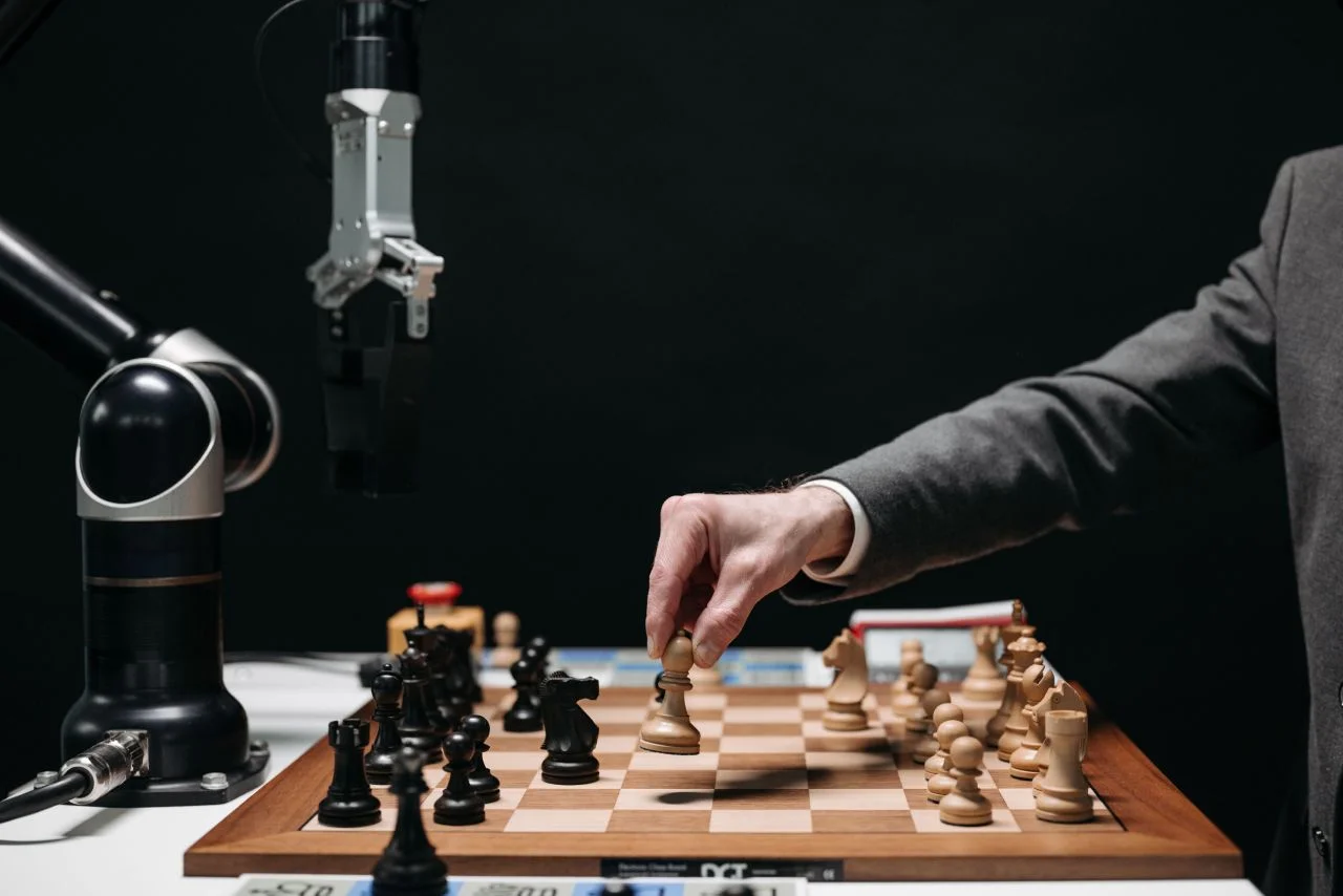 A person in a suit moves a chess piece on a chessboard while a robotic arm hovers nearby, possibly playing or observing the game. The background is dark and focused on the chess match.